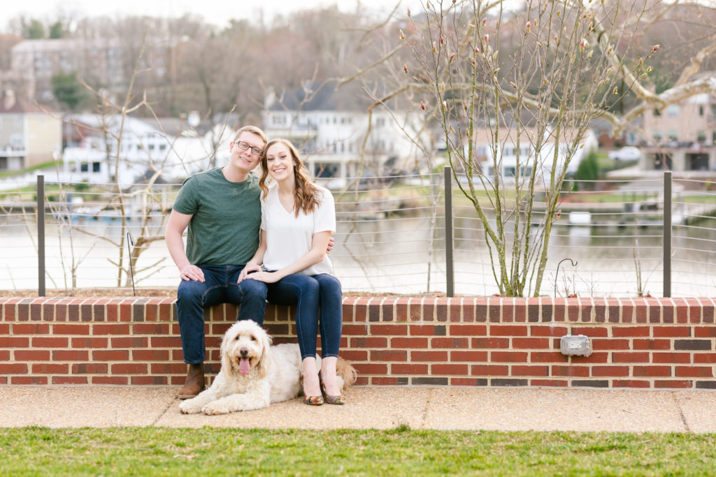 couple in casual clothes sitting with on brick ledge with dog laying at their feet, all smiling looking at camera
