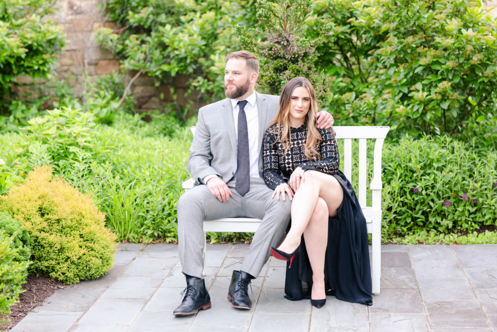Couple sitting on bench in garden. Girl with legs crossed, in Louboutin red bottom heels and black dress. Guy in grey suit