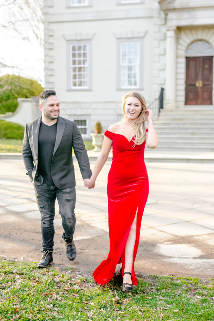 Blonde girl in red dress walking in front of guy in all black, pulling him towards her as she looks at camera
