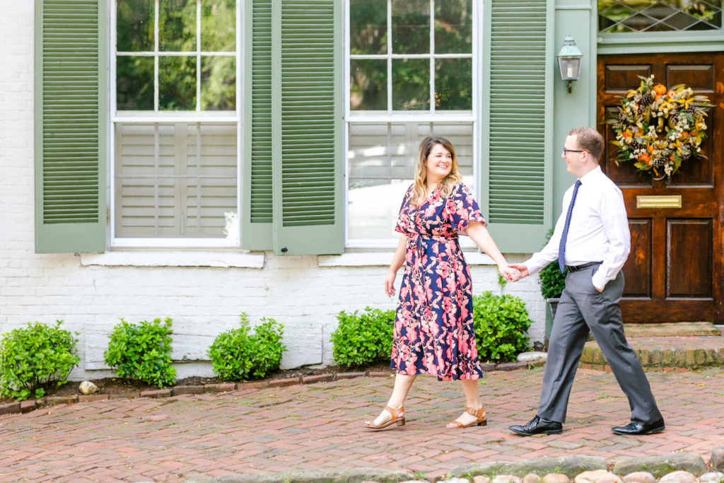Couple walking hand in hand down brick paved street with green and white shutters behind them. Girl in pink and blue floral dress, guy in dress pants and white shirt