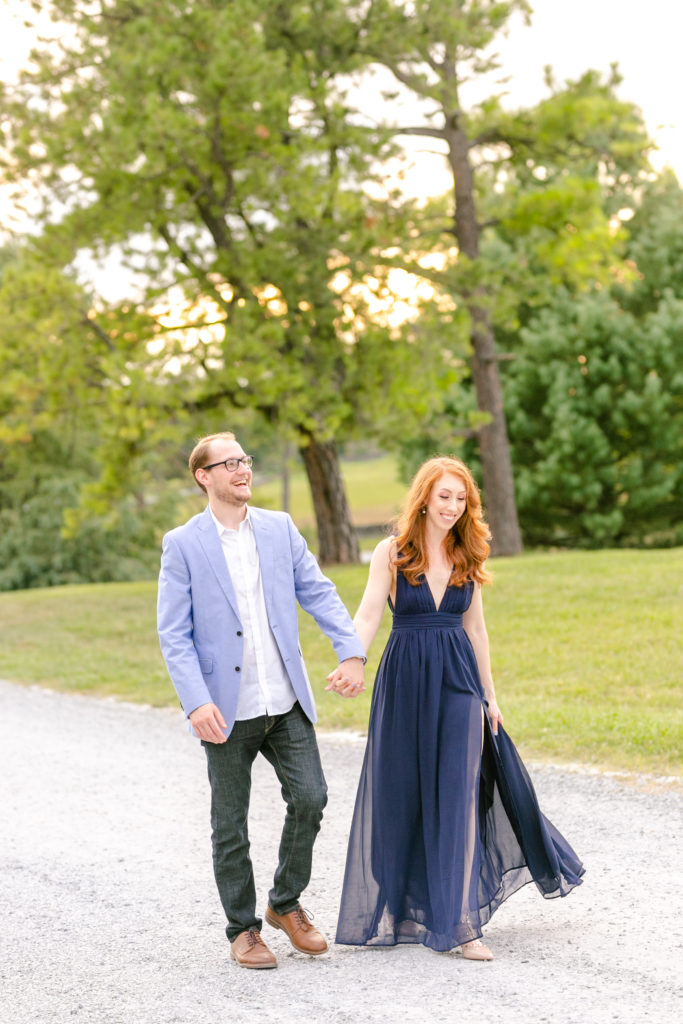 Red head girl in long blue dress walking hand in hand with guy down path at sunset - they're laughing