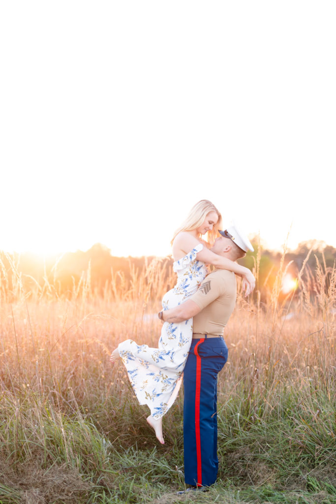 Girl in white and blue floral dress being lifted by a Marine in a field of tall grass at sunset