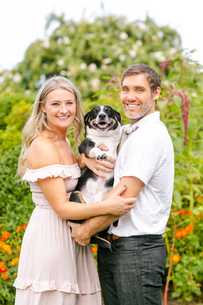 blonde girl in pink dress and guy in white shirt smiling as they hold their beagle mix dog in between them in a garden