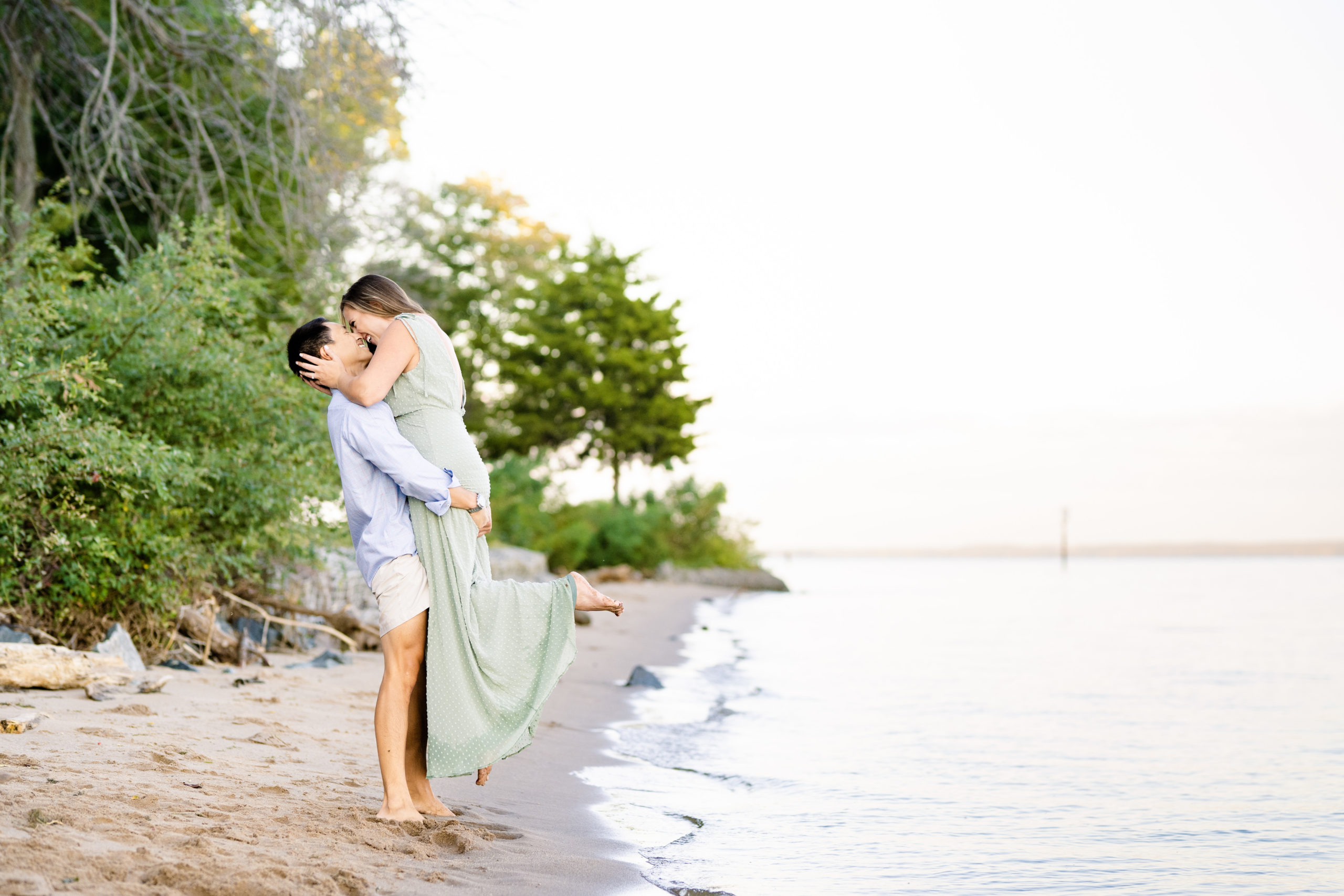 guy in tan shorts and blue shirt lifting girl in mint green dress while they laugh and kiss on the beach